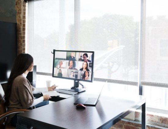 A group of creative professionals meet virtually to discuss and brainstorm ideas. A young female creative professional is working in an office while talking with colleagues who are telecommuting from their homes.